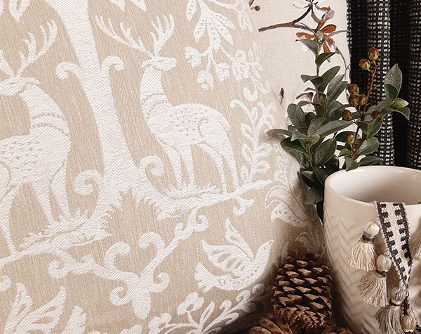 A pillow featuring fabric with a pattern of deer and trees in light neutrals next to holiday greenery and tassel trim.