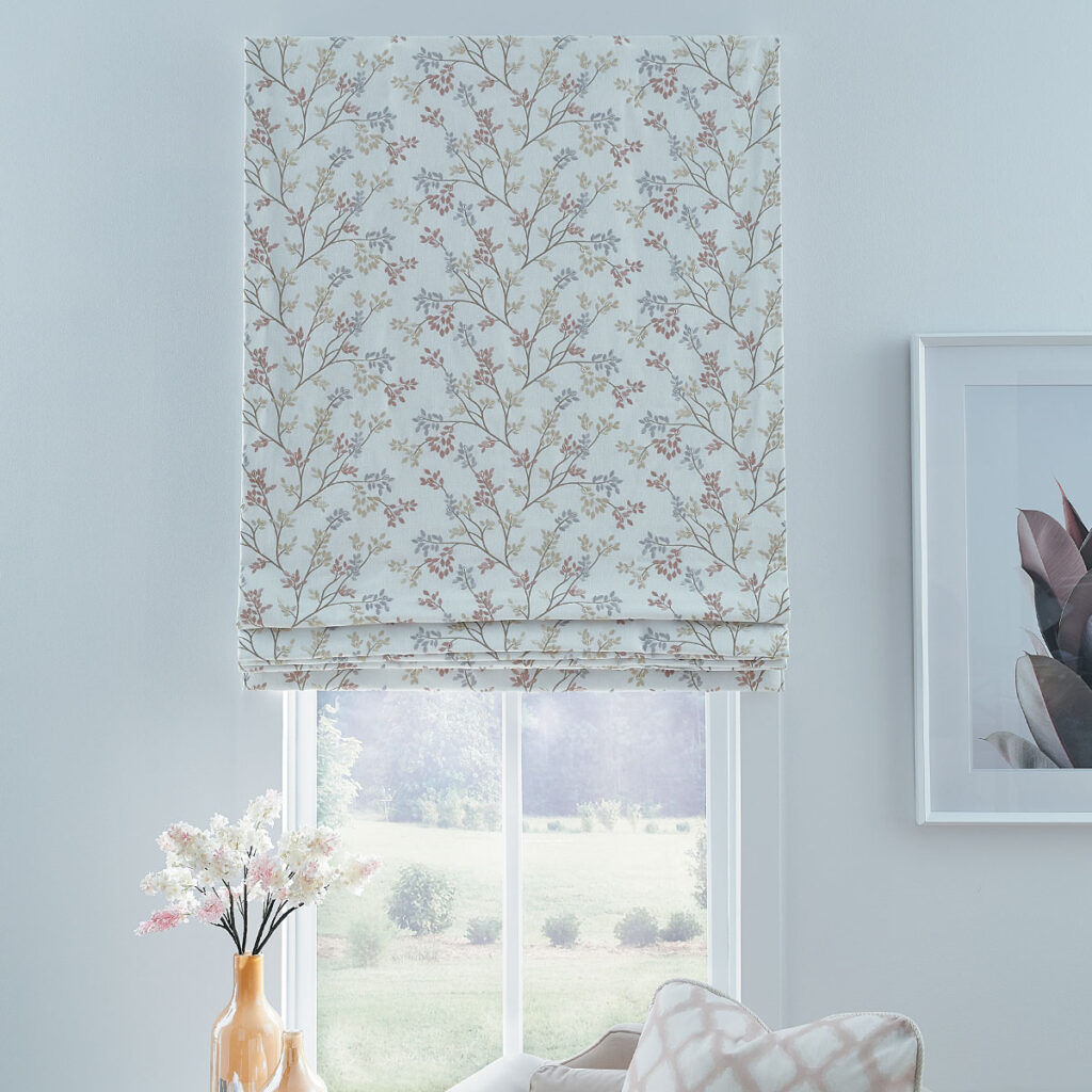 A Flat Roman Shade covered in delicate floral embroidery in a sitting area with white walls and decor in pastel pink and peach.