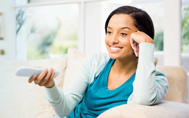 Woman on couch holds Amp Motorization remote control, pointed at window treatments off camera.