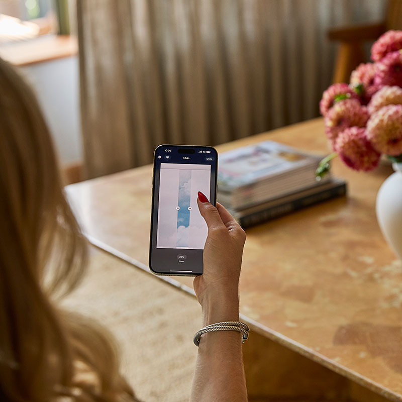 Looking over the shoulder of a woman in a living room holding a smart phone with the PowerView App on the screen.