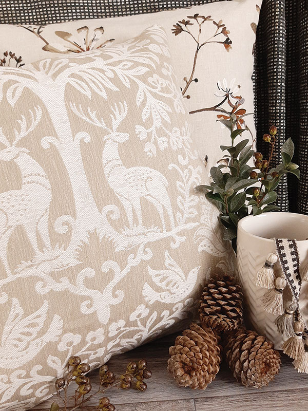 A pillow featuring fabric with a pattern of deer and trees in light neutrals next to holiday greenery and tassel trim.