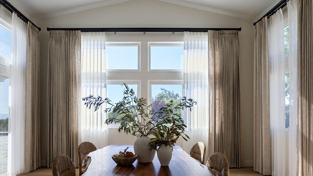 A dining room with windows on all sides that feature neutral sheers and drapery, with a rustic table in the center.